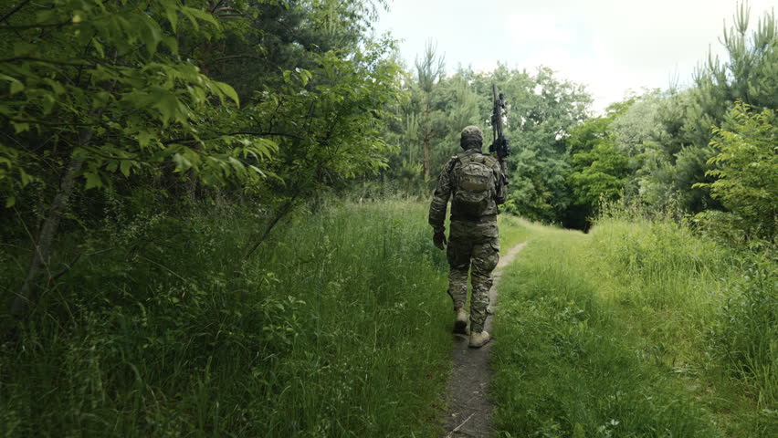 Legs of a camouflaged soldier in the middle of a footpath in a field of grass on a mission. Walking and aiming a AR-15 carbine rifle with a red laser collimator and a magnifier