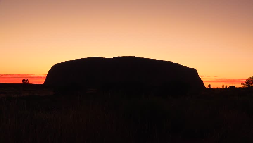 Iconic Uluru monolith silhouetted against a fiery sunset. Dramatic orange and red sky illuminates desert plains. Natural wonder at dusk.