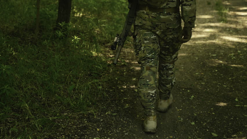 Legs of a camouflaged soldier in the middle of a footpath in a field of grass on a mission. Walking with a AR-15 carbine rifle with a red laser collimator and a magnifier attached to his backpack