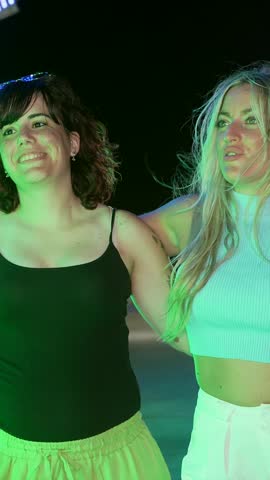 Two young women laughing and having fun together on a colorful ride at an amusement park at night, illuminated by vibrant green, blue, and pink neon lights during a summer evening festival