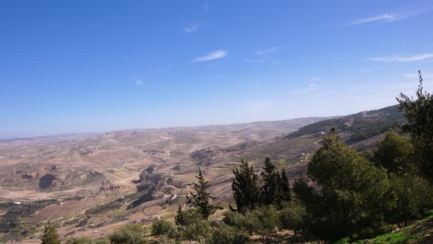 A wide panoramic view of the arid desert landscape and rolling hills as seen from the Mount Nebo lookout point in Madaba, Jordan under a clear blue sky.