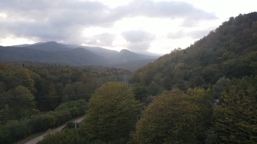 Aerial drone footage capturing a smooth flight over vibrant autumn mountain forests. The composition reveals a scenic valley with a small house nestled among the trees, with distant mountain ranges under a cloudy sky
