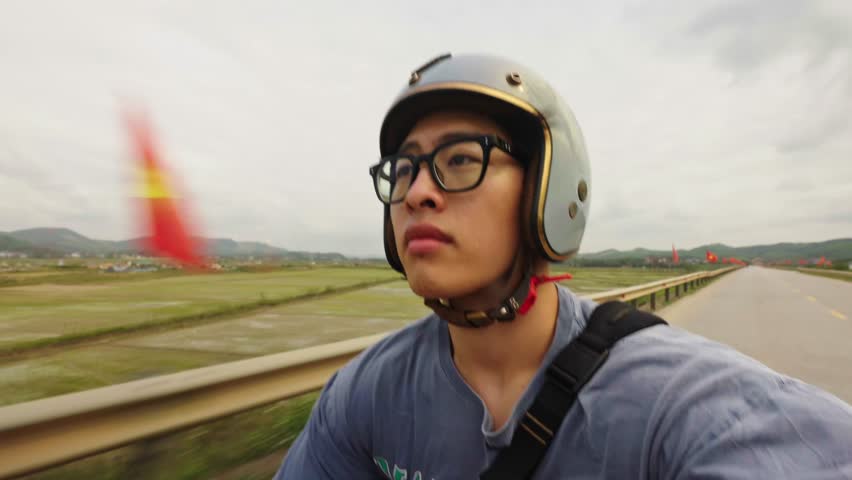 Young vietnamese man taking selfie while riding scooter with national flag on road trip through rural Vietnam, freedom and adventure on vacation