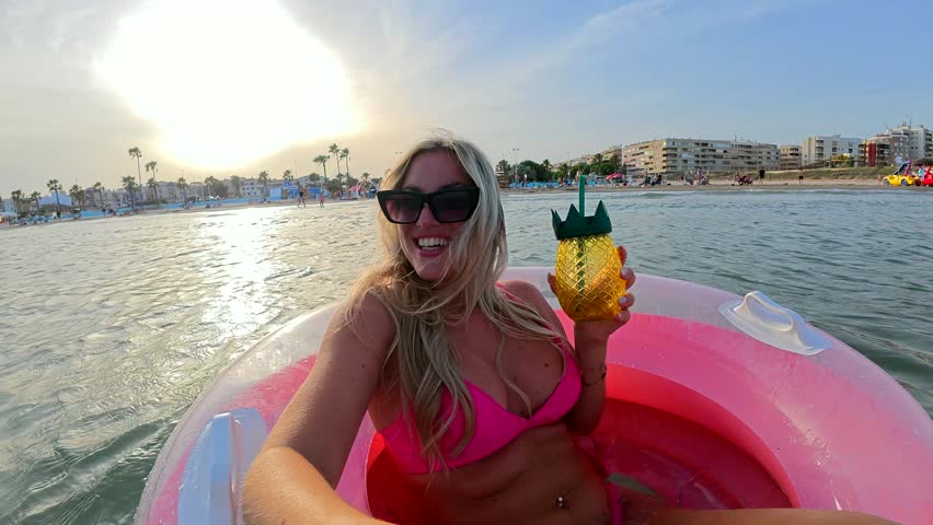 Happy young woman in a pink bikini floating on an inflatable ring at sea, holding a pineapple shaped cocktail and enjoying her summer vacation at sunset while smiling at the camera