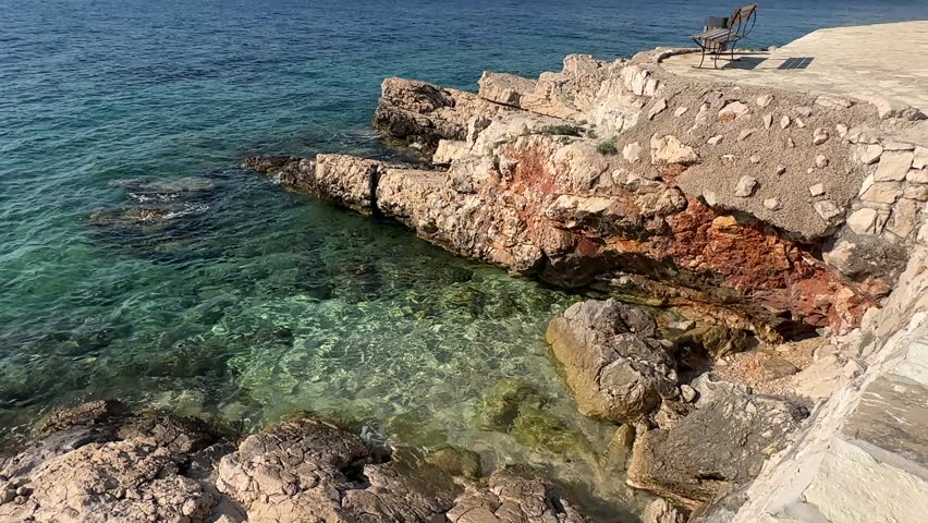 People relax by the rocky shore where clear water meets the land. A chair is placed nearby for sitting. Waves gently touch the rocks under the bright sun.