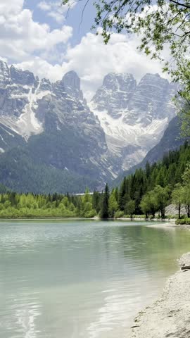 "Scenic view of a turquoise alpine lake with a lush green pine forest and majestic rocky mountain peaks covered in snow under a blue cloudy sky"