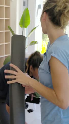 Vertical video: Exchanging mats, two women preparing for yoga at home, light-blue-top woman walking. Livingroom, lifestyle, minimalist, modern, fitness, interior, vertical video