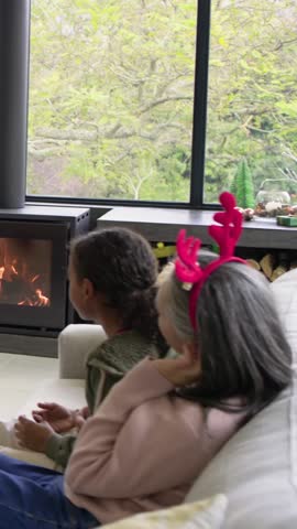 Vertical video: Fire drawing attention, woman in red antlers turning green jar to child at home. Cozy, hearth, family, rustic, festive, logs, vertical video