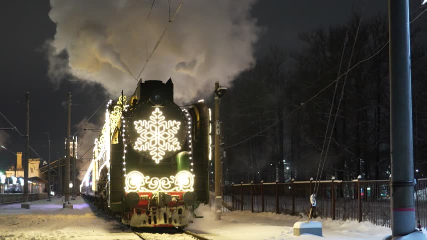 A festive steam locomotive, brightly adorned with a large snowflake light display, moves along snow-covered tracks at a station during a dark winter night.