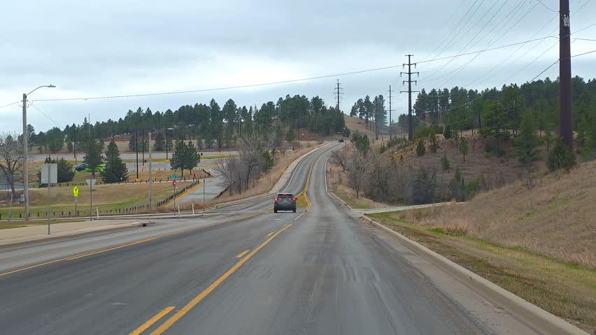 United States, April 2 2026: Curving road in Rapid City South Dakota, cars pass pine covered hills and power lines under a cloudy sky in a travel video frame