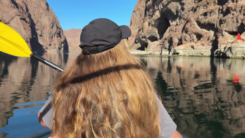 Kayaking through a rocky Colorado River canyon, a young paddler moves across calm water with steep desert cliffs reflected in the clear surface