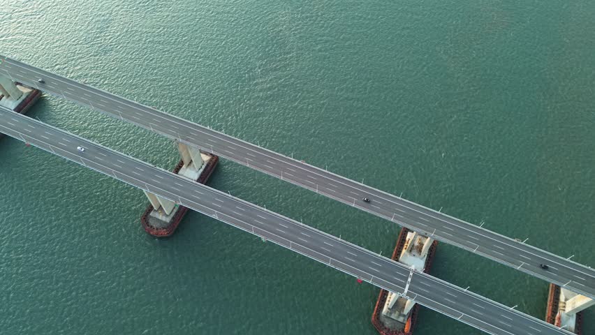 Artistic vertical overhead top-down sliding aerial clip captured in gentle pre-sunset golden dusk above twin diagonally offset parallel cross-sea highway bridge at Pingtan Strait, Fuzhou. Minimalist diagonal symmetrical grid geometric layout formed by two straight offset concrete road decks, evenly spaced paired rust-red pier fenders and linear bridge shadows against smooth emerald rippling ocean. Tiny moving sedans and a green freight truck add subtle dynamic layers, frameless pure marine abstract shot with extra landscape removed.