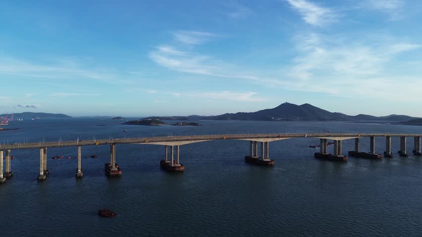 Wide horizontal aerial drone tracking footage shot before sunset over Pingtan Strait Highway Bridge, Fuzhou Fujian, China. Panoramic eye-level aerial shot captures twin six-lane cross-sea viaduct stretching across deep blue bay water. Small fishing boats scatter on sea surface, layered green offshore islets and mountain silhouettes line the horizon, red port cranes appear on far left shore, wispy cirrus clouds fill bright clear blue sky.