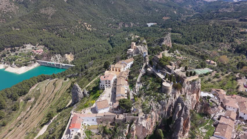 Drone shot of Guadalest old town on rocky cliffs above a blue reservoir.