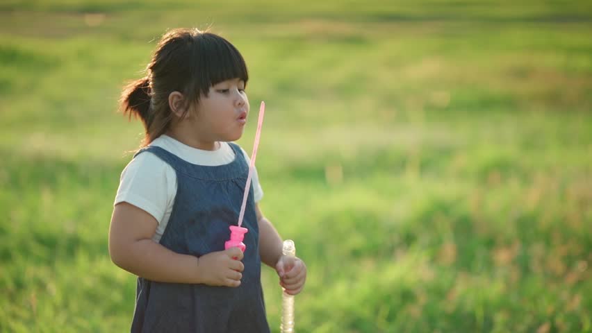 Adorable Asian girl holding a bubble wand and watching floating soap bubbles in slow motion outdoors. Happy childhood, playful moments, summer lifestyle, freedom, and innocence concept.