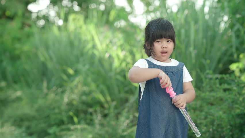 Adorable Asian girl holding a bubble wand and playing outdoors in a green public park. Happy childhood, outdoor activity, summer fun, innocence, and carefree lifestyle concept.