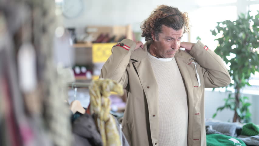 Interested man standing in modern clothing shop and trying new beige raincoat for cold season 