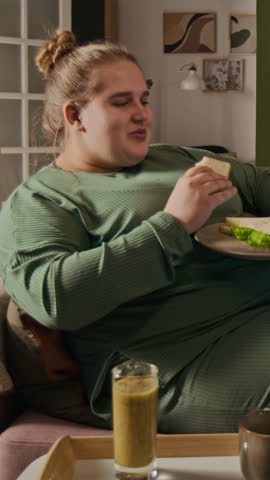 Vertical video of plus-size Caucasian young adult woman sitting on sofa and eating sandwich. Clip is ideal for body positivity campaigns or intuitive eating concepts