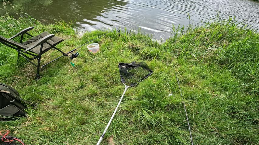 Peaceful fishing spot on river bank with chair, landing net, rod and bait bucket, outdoor leisure hobby