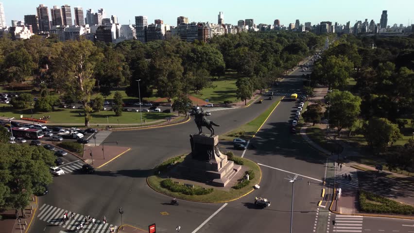 Aerial footage of the Monument to Justo José de Urquiza located in the center of a major traffic circle within Parque Tres de Febrero in Buenos Aires, Argentina. The camera gradually approaches the historic equestrian monument while revealing busy intersections, tree-lined parkland, surrounding avenues, and the modern skyline of the Argentine capital. Bright afternoon light highlights the contrast between green urban spaces and dense city development, making the clip suitable for travel, tourism, transportation, infrastructure, city planning, history, architecture, and South American destination projects.