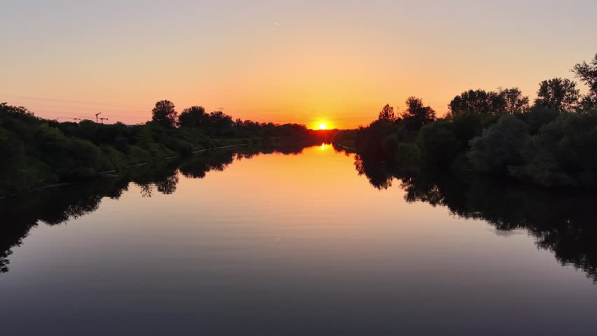 Aerial view of a tranquil river at sunset, showcasing vibrant orange and yellow hues reflecting on the water, surrounded by lush green trees and serene landscape