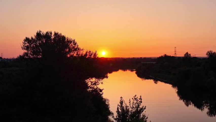Sunset over a tranquil river with lush greenery and silhouettes of trees, showcasing a gradual transition of colors in the sky as the sun descends behind the horizon