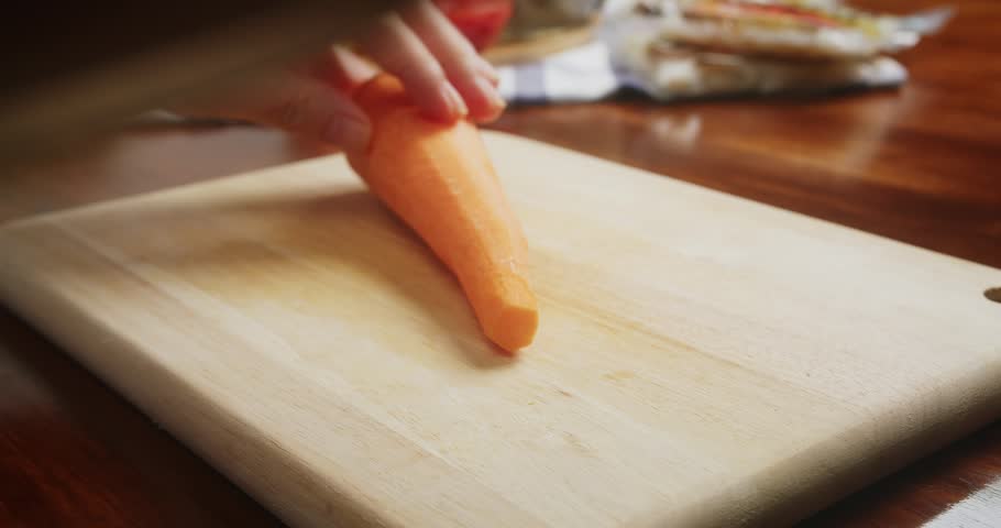 Close up of chef hands slicing fresh carrots on a wooden cutting board with a sharp kitchen knife. Fresh carrots are cut into diagonal pieces during food preparation in a home kitchen.