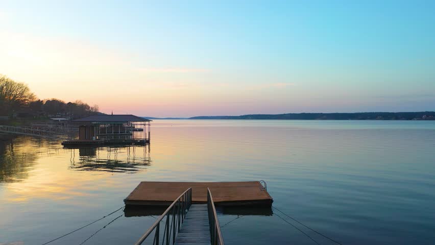 Sunset drone shot over Dale Hollow Lake, Tennessee, with residential shoreline, private boat houses, wooded hills, calm reservoir water, and soft golden reflections in peaceful dusk light.