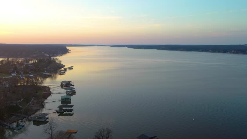 Peaceful Lake of the Ozarks drone view with waterfront homes, covered boat docks, forested coves, calm evening water, and warm sunset glow across the scenic Missouri lake landscape at dusk.