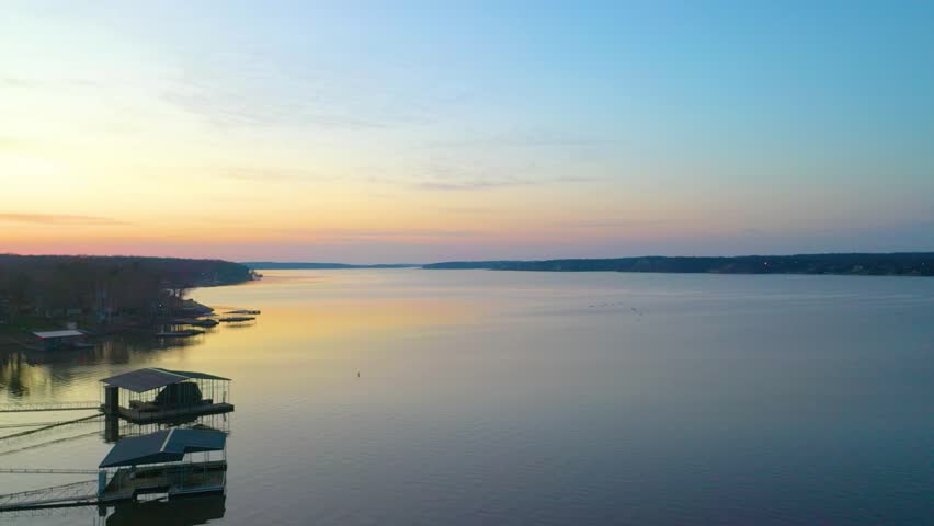 High drone view above Beaver Lake at dusk, looking across calm water, wooded Arkansas shoreline, docked boat houses, lakefront homes, and soft sunset colors over the reservoir.