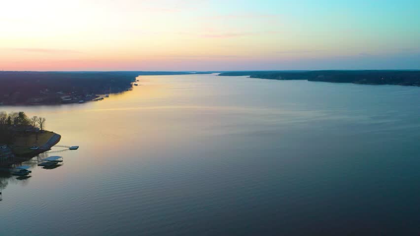 Wide aerial view of Bull Shoals Lake, showing quiet docks, lakeside homes, tree-covered Ozark shores, pastel sunset light, and calm open water stretching toward the horizon.