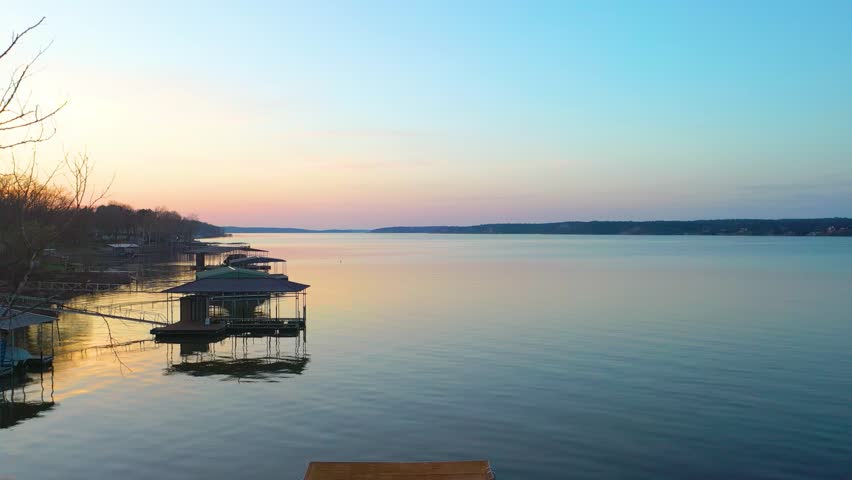 High drone view above Lake of the Ozarks at dusk, looking across calm water, wooded Missouri shoreline, docked boat houses, lakefront homes, and soft sunset colors over the wide reservoir.