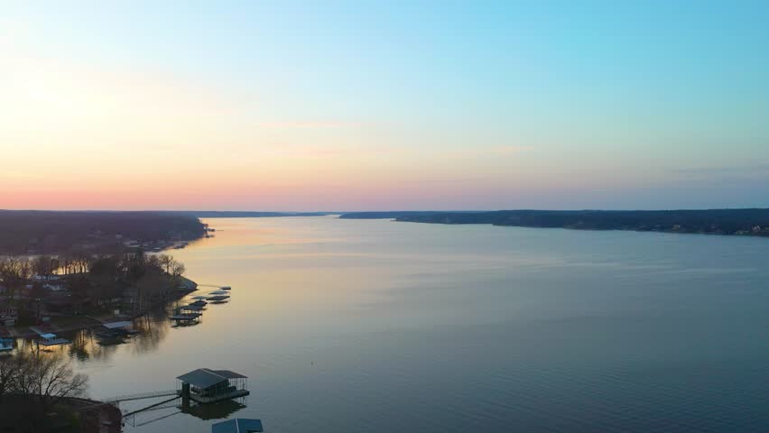 High drone view above Norris Lake at dusk, looking across calm water, wooded Tennessee shoreline, docked boat houses, lakefront homes, and soft sunset colors over the reservoir.