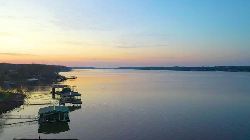 Drone aerial over Beaver Lake at sunset, with calm Arkansas water, wooded Ozark shoreline, lake houses, covered boat docks, pastel sky, and golden reflections across the wide reservoir.
