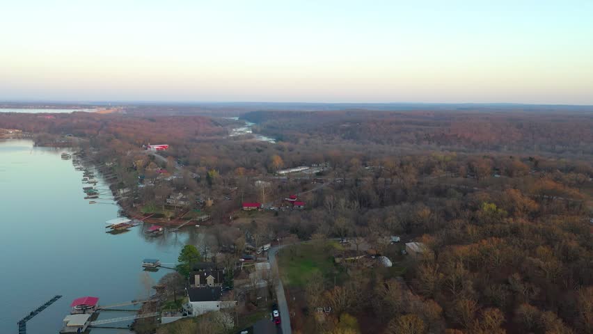 Drone aerial approaches a lakefront vacation house with large deck, screened porch, wooded yard, nearby homes, and calm water views along a quiet residential shoreline at sunset.