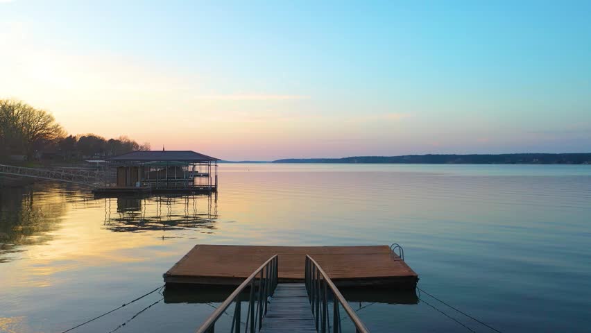 High drone view above Table Rock Lake at dusk, looking across calm water, wooded Missouri shoreline, docked boat houses, lakefront homes, and soft sunset colors over the reservoir.