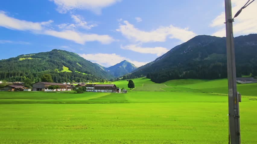 Austria rail travel through a peaceful mountain valley, showing bright meadows, chalet style homes, roadside trees, and forested peaks under summer skies.