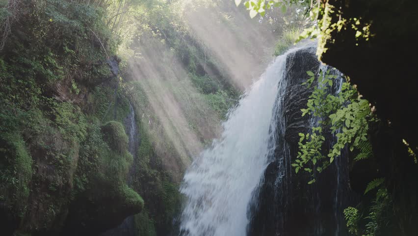 Beautiful waterfall with sun rays in a lush green forest in Oita. A mystical video of Ameushi Falls in Taketa, Oita, Japan, with dramatic sunbeams (crepuscular rays) piercing through the trees. The powerful waterfall, surrounded by moss-covered rocks and fresh greenery, creates a serene and therapeutic atmosphere. Perfect for nature, travel, wellness, and healing concepts. with natural sound