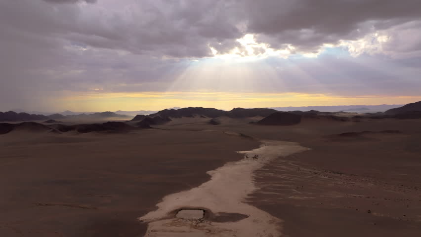 Cinematic aerial as crepuscular sun rays break through clouds over the dunes and dry riverbed of the Namib desert at dawn. Soft light wraps the ancient dunes in a calm mood.