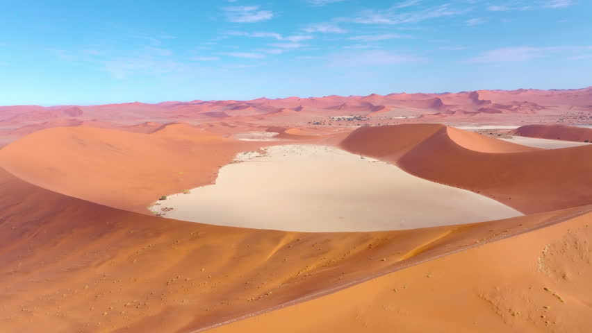 Aerial over the immense red dunes and pale pan of Sossusvlei under a clear blue sky. The towering red dunes glow against the pale cracked clay pans of this ancient desert wilderness.