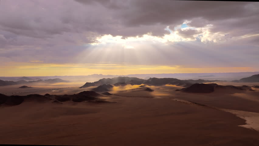 Aerial as sunbeams sweep over the misty mountains and dunes of the Namib desert at first light. Soft light and drifting mist wrap the ancient dunes and mountains in a calm cinematic atmosphere.