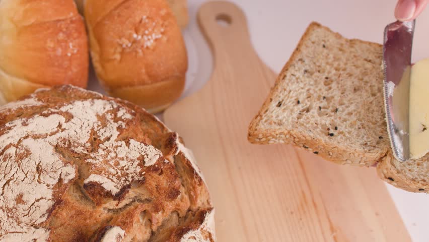 Close-up shot of a knife spreading butter on multigrain bread surrounded by artisanal loaves.