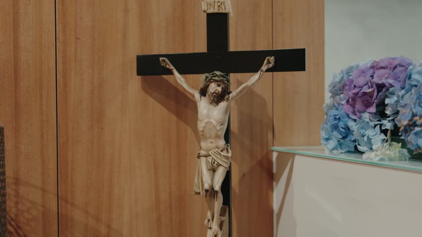 Carved crucifix of Jesus Christ on a black cross beside blue hydrangeas on a wooden church wall