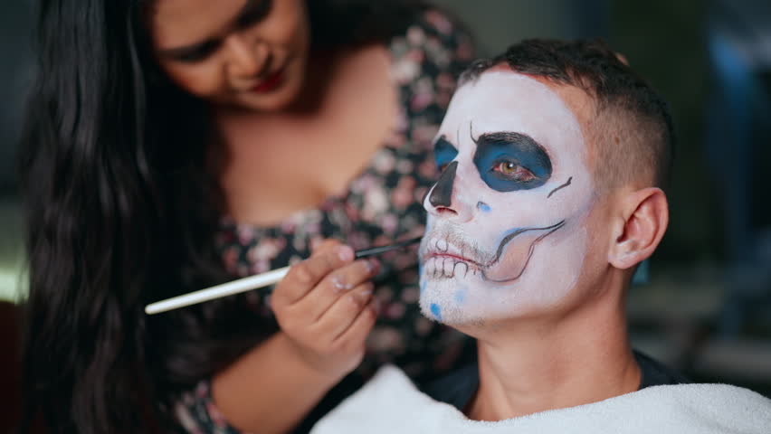 Woman carefully applies traditional Day of Dead sugar skull makeup on young man using fine brush. White face paint with black and blue details around eyes, painted teeth and nose create haunting look.