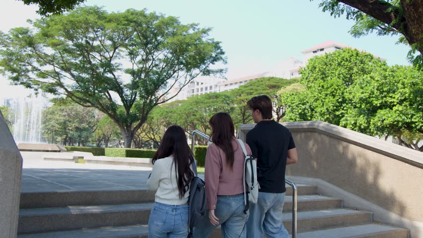 Three young adults with backpacks walk up concrete stairs in a sunny university park.
