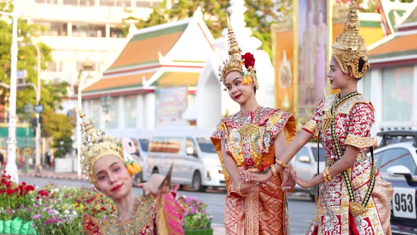 Three female dancers in ornate traditional costumes perform graceful hand movements on a city street.