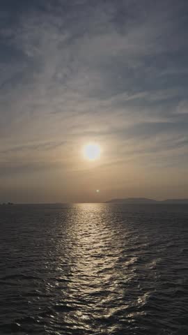 Static wide shot of a hazy golden sunset over the calm ocean horizon, with sunlight reflecting on the rippling water surface under a cloudy sky.