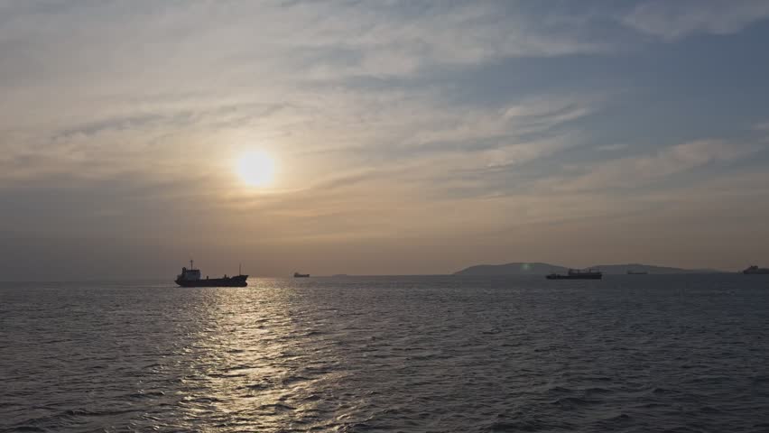 Static wide shot of a large cargo ship silhouette sailing on the calm ocean during a hazy golden sunset, with sunlight reflecting on the water surface.