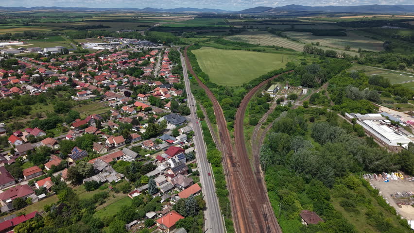 An expansive aerial view showcases a Hungarian village nestled beside a complex railway junction and lush green agricultural fields.