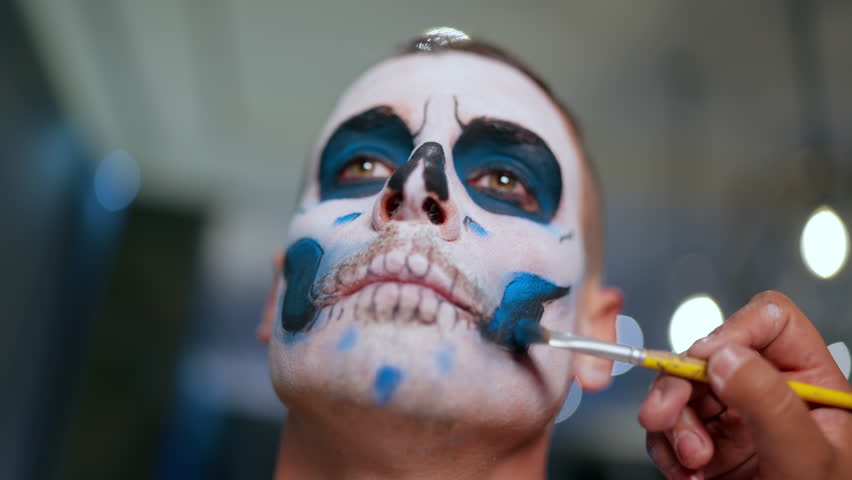 Makeup artist carefully applies intricate Day of the Dead sugar skull face paint on man using yellow brush. Close-up shows detailed white, black and blue design with bokeh lights in background.